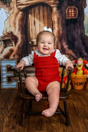 Smiling baby in red romper sits in wooden chair with Winnie the Pooh backdrop and Tigger plush toy.