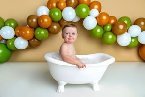 Happy baby sitting in a white toy bathtub with bubbles, surrounded by green, white, and brown balloon arch.