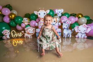 Baby girl sits on chair at safari-themed first birthday party with balloon arch and animal cutouts.