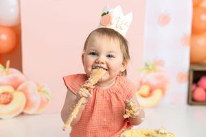 Baby girl in peach dress and crown smashes first birthday cake against peach balloon backdrop.
