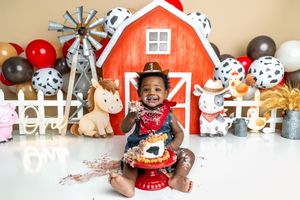 Baby smiling during farm-themed cake smash with red barn backdrop, cow print balloons, and toy animals.
