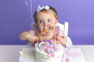 Baby girl in purple tutu celebrates first birthday smashing a unicorn cake, wearing beads and a bow.