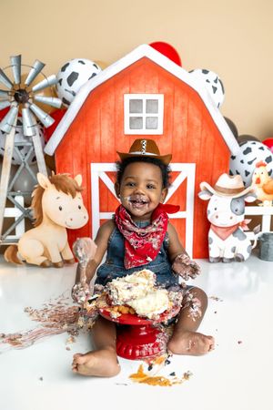Smiling baby in cowboy hat enjoys cake smash with farm-themed backdrop featuring red barn, windmill, and animal cutouts.