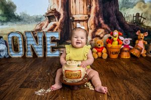 Baby girl in yellow outfit smiles at Winnie the Pooh themed first birthday cake smash with honey pot and plush toys.