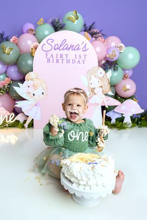 Baby girl in green dress smashes cake at Solana's Fairy 1st Birthday party with balloon backdrop.