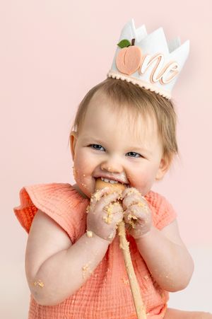 Baby girl in peach dress and “one” crown smiles while eating cake during first birthday smash cake photo session.