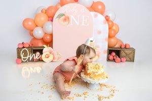 Baby girl in peach outfit smashes first birthday cake at ONE-themed party with balloon arch backdrop.