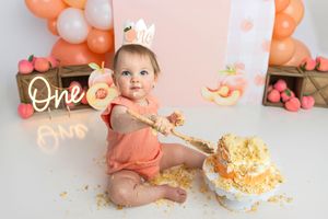 Baby girl in peach romper smashes first birthday cake surrounded by peach balloons and One signs.