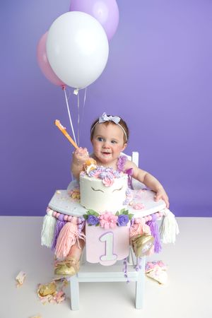 Baby girl celebrating her first birthday with a unicorn cake, balloons, and a sparkler in a purple-themed photo shoot.