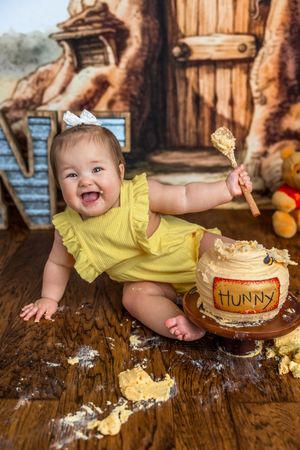 Happy baby girl in yellow romper smashing a Winnie the Pooh Hunny pot cake during first birthday photoshoot.