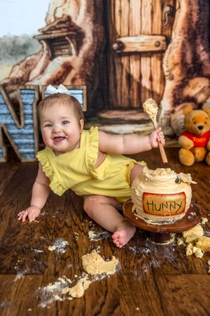 Baby girl in yellow romper smashes Winnie the Pooh Hunny pot cake on wooden floor at first birthday photo shoot.