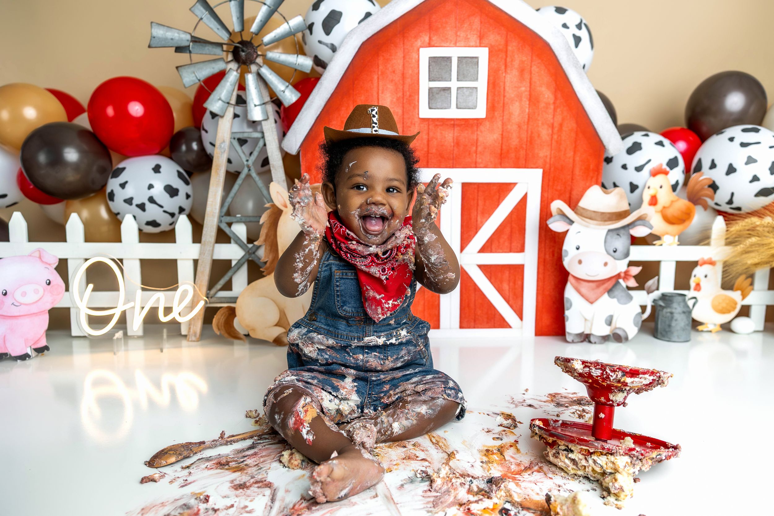 Smiling baby in cowboy hat and bandana enjoys a farm-themed first birthday cake smash with red barn backdrop.