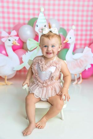 Smiling baby girl in pink ruffle romper sits on white chair at first birthday party with swan decorations and balloons.
