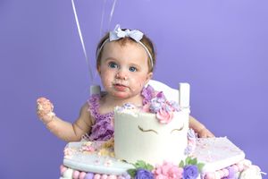 Baby girl in purple romper smashes unicorn birthday cake, face covered in frosting, purple background.