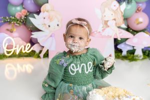 Baby girl in green dress smashes first birthday cake surrounded by fairy-themed decorations and balloon backdrop.