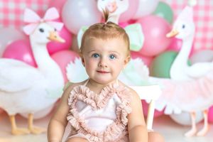 Adorable baby girl with blue eyes in pink ruffle outfit at swan-themed first birthday party with balloons.