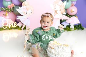 Baby girl in green “one” sweater smashes first birthday cake surrounded by fairy-themed balloon decorations.