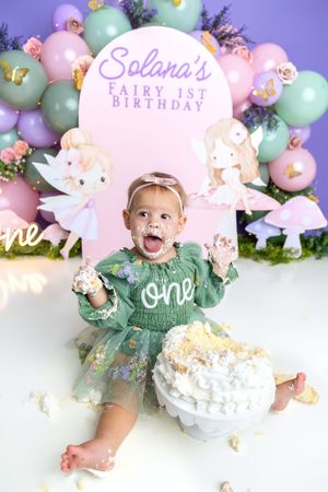 Baby girl in green tutu smashes cake at Solana's Fairy 1st Birthday party with balloon backdrop.