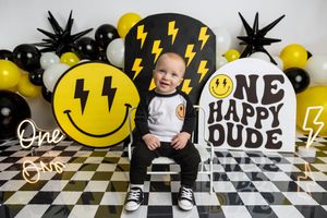Baby boy's first birthday photo with smiley face balloons, lightning bolt props, and One Happy Dude sign.