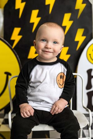 Smiling baby in black and white raglan shirt with smiley face, posed before lightning bolt backdrop.