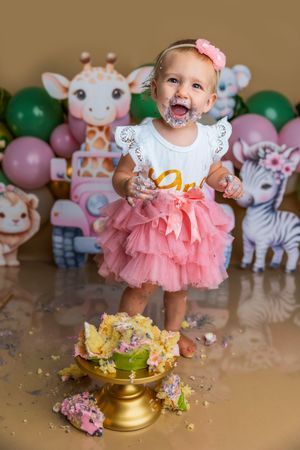 Baby girl in pink tutu smashes first birthday cake surrounded by safari animal decorations and balloons.