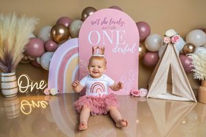 Baby girl in pink tutu sits before a rainbow ONE-derful backdrop with balloons for her first birthday cake smash photo.