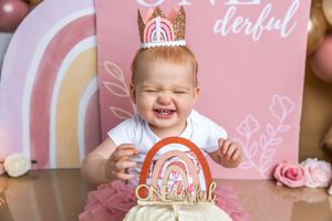 A joyful baby girl in a pink tutu and crown celebrates her first birthday with a rainbow cake smash.