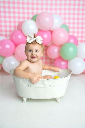 Smiling baby girl in white bathtub with rubber duck, pink gingham backdrop and colorful balloons.
