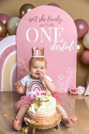 Baby girl in pink tutu smashes first birthday cake under ONEderful sign with balloon garland backdrop.