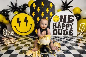 Baby's first birthday cake smash with smiley face and lightning bolt decorations, black and yellow balloon backdrop.