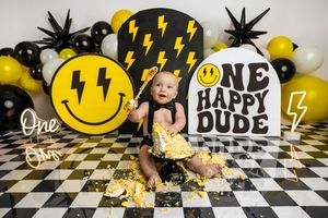 Baby's first birthday cake smash with yellow smiley face and lightning bolt decorations on checkered floor.