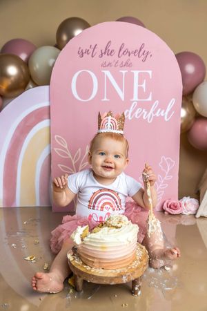 Baby girl in pink tutu and crown smiles at first birthday cake smash with ONEderful backdrop and balloon garland.