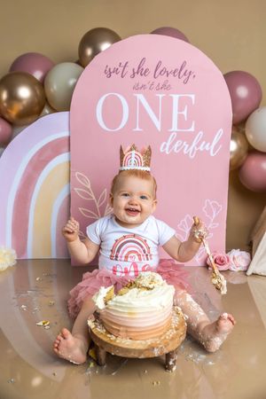 Baby girl in pink tutu smashes first birthday cake in front of ONEderful balloon backdrop with rainbow decorations.