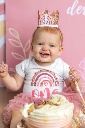 Smiling baby girl in pink tutu and glitter crown celebrates first birthday with smash cake.