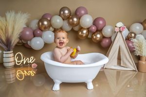 Baby smiling in white bathtub with rubber duck, surrounded by pink gold white balloons and One neon sign decor.