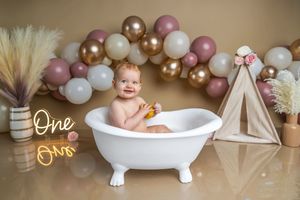 Baby smiling in white bathtub with gold pink balloons and teepee decor for first birthday photo shoot.