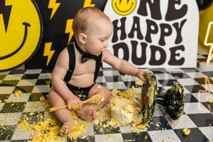 Baby boy smashing black and gold cake at One Happy Dude first birthday party on checkered floor.