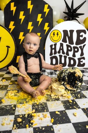 Baby boy cake smash photo with smiley face and lightning bolt backdrop, black suspenders, smashed yellow and black cake.