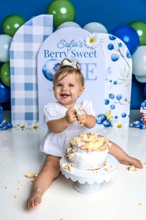 Baby girl smashes first birthday cake at Berry Sweet One blueberry themed party with balloon backdrop.