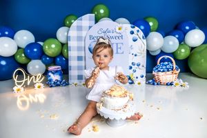 Baby girl celebrates first birthday smashing cake at blueberry themed party with blue and green balloon backdrop.