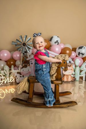 Baby girl in denim overalls smiles on wooden rocking horse at farm-themed first birthday party with balloons.