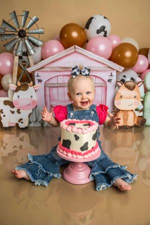 Baby girl in denim overalls smiles at farm-themed first birthday cake smash with cow print cake and balloon backdrop.