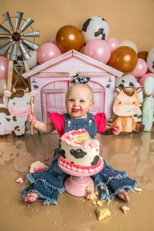 Baby girl in denim overalls smashes cow-print birthday cake at farm-themed first birthday party with pink barn backdrop.