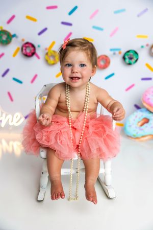 Smiling baby girl in pink tutu skirt and pearl necklace sitting on white chair with colorful donut birthday backdrop.