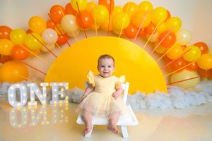 Baby girl in yellow dress sits before colorful orange balloon arch with ONE sign at first birthday photo shoot.