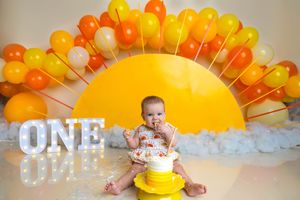 Baby enjoys first birthday cake smash with sunny balloon arch backdrop and glowing ONE marquee letters.