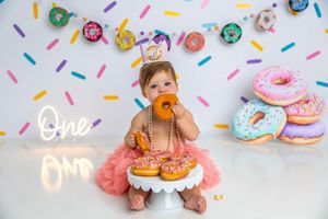 Baby girl in pink tutu celebrates first birthday with donuts and colorful sprinkle backdrop at a donut-themed cake smash.