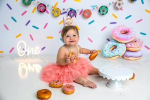 Baby girl in pink tutu sits on floor eating donuts at colorful sprinkle-themed first birthday cake smash.