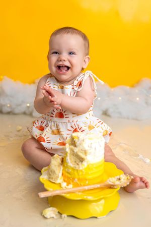 Happy baby girl clapping hands at first birthday cake smash session with yellow backdrop and sunshine outfit.