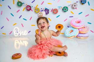 Baby girl in pink tutu celebrates first birthday with donuts and colorful sprinkle backdrop and neon One sign.